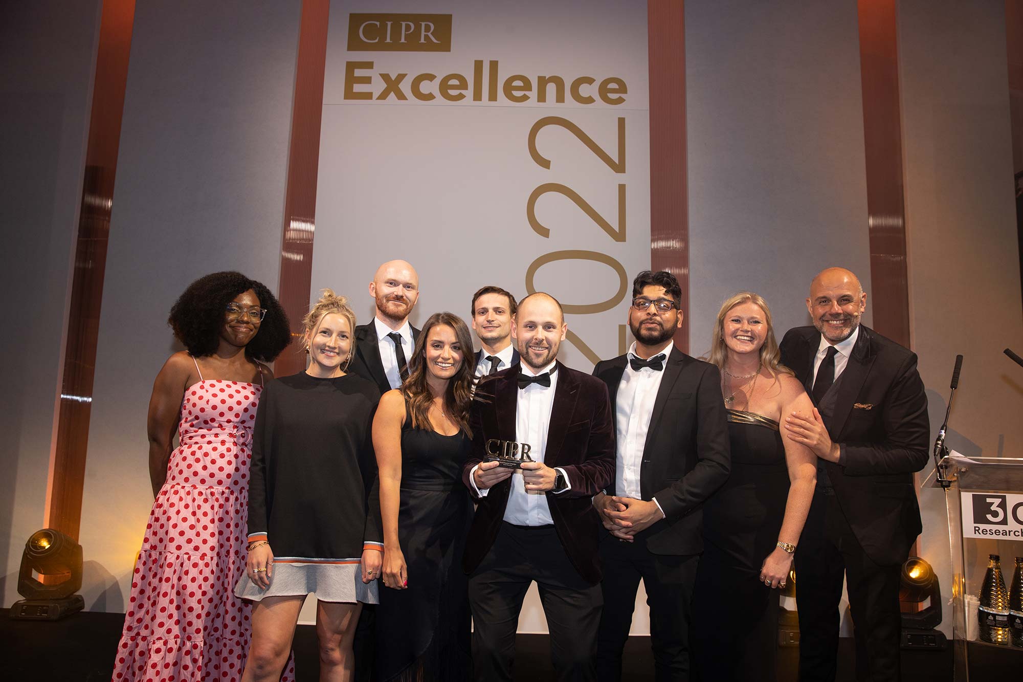 Photo of a group of CIPR 2022 Excellence Awards winners on stage, holding up their trophy, pictured with the events ceremony MC, Jason Mohammed