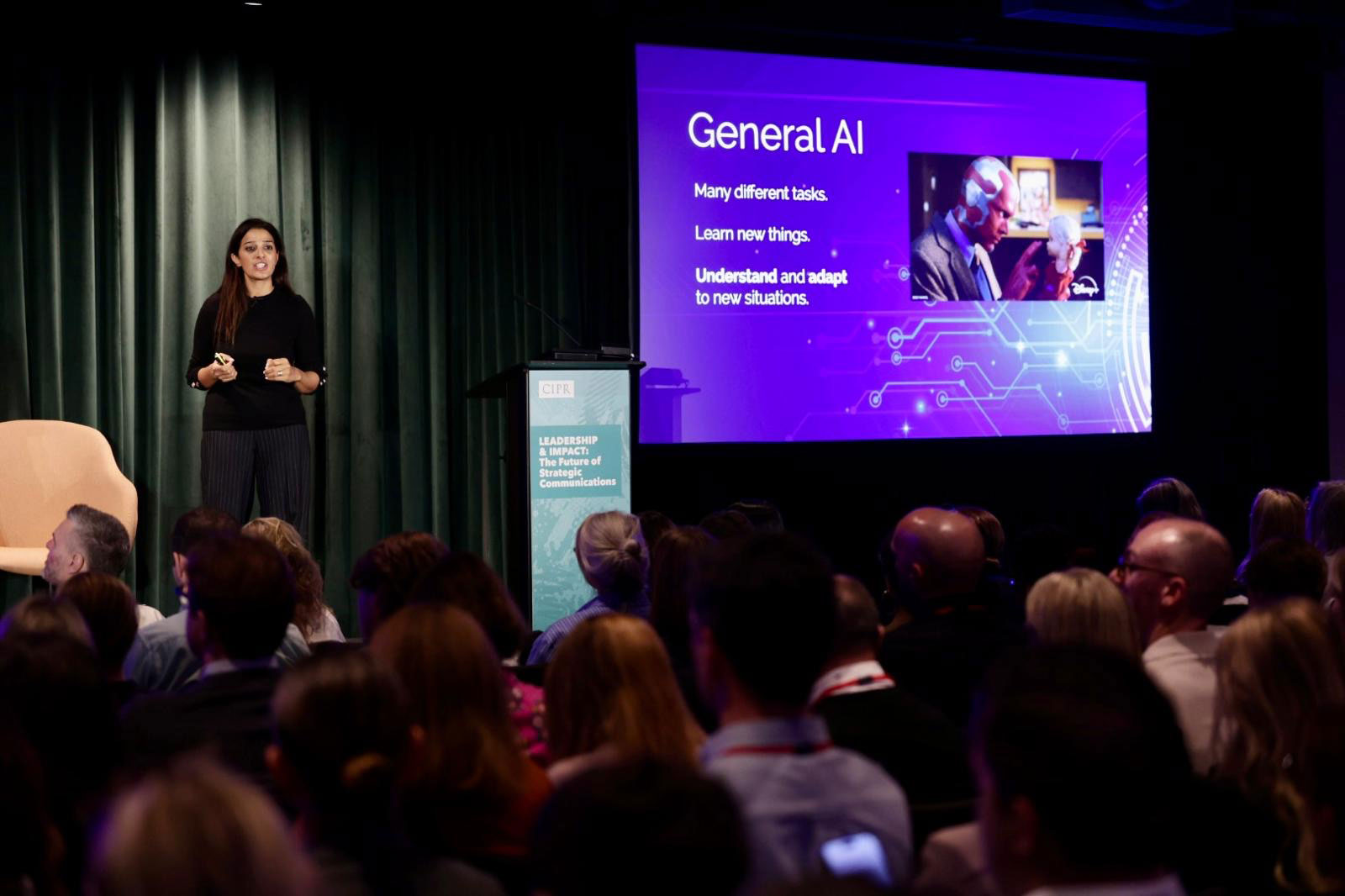 A speaker on stage at the CIPR 2025 Leadership and Impact annual conference, presenting a slide on AI to a seated audience.