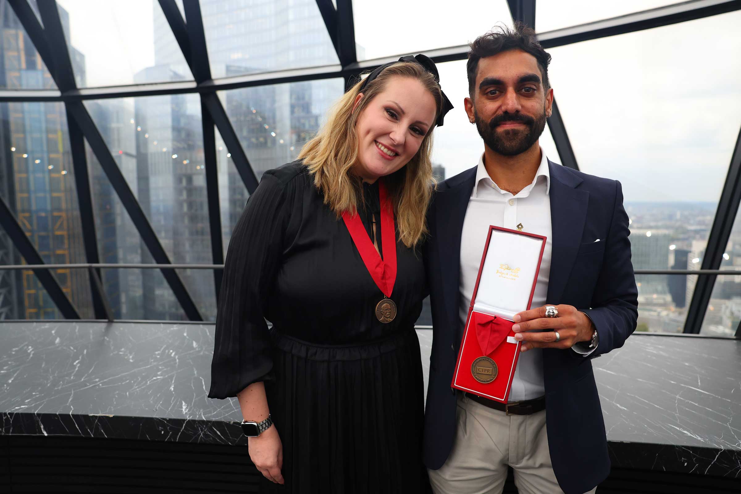 Anne-Marie Lacey, wearing the Sir Stephen Tallents' Medal, and Sheeraz Gulsher, holding the President's Medal, at the CIPR Annual Fellows' Lunch at the Gherkin in London, with the City skyline behind them.